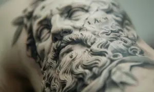 Detalle de un tatuaje de microrealismo conceptual de un rostro clásico con barba rizada y corona de laurel.