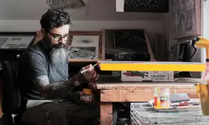 Thomas Hooper, con barba y gafas verdes, pinta un objeto amarillo en su estudio.
