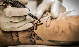 Manos enguantadas crean un diseño de letra con una máquina de tatuar. Un tatuaje con buena tinta se está formando en la piel.