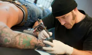 Tatuador con gorro negro y guantes blancos tatuando el brazo de un cliente con tatuajes coloridos.