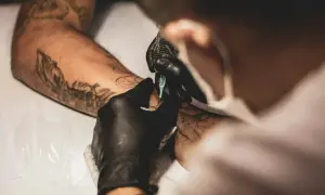 Artista con guantes negros usa máquina para crear un tatuaje en un brazo con diseños previos. La emla para tatuajes ayuda a que el proceso sea más cómodo.
