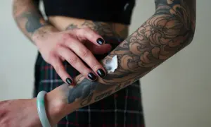 Mano con uñas negras aplicando crema sobre un tatuaje de estilo dark work en el brazo, con diseño de olas y flores.