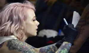 Artista de tatuajes con cabello rosa, con un diseño de megan massacre en su brazo, mientras otro artista trabaja en su piel.