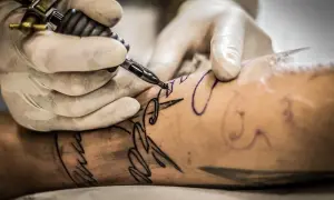 Manos enguantadas trabajan con una máquina de tatuar sobre un brazo, delineando un diseño. Aprende como cuidar un tatuaje recien hecho.