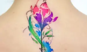 Un vibrante tatuaje de flor estilo acuarela en la espalda, con tonos rosas, morados y verdes. Un diseño de tatuajes de colores para mujer que evoca la naturaleza.