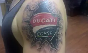 Tatuaje de un logo de Ducati Corse en el hombro, con efecto de piel rasgada.