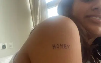 Un delicado tatuaje handpoke con la palabra "HONEY" en el brazo de una mujer sonriente.