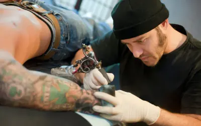 Tatuador con gorro negro y guantes blancos tatuando el brazo de un cliente con tatuajes coloridos.