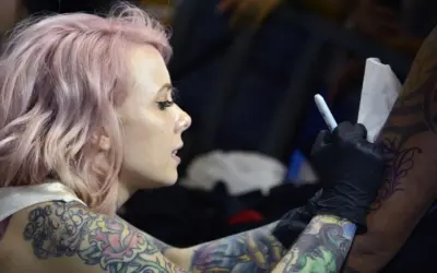 Artista de tatuajes con cabello rosa, con un diseño de megan massacre en su brazo, mientras otro artista trabaja en su piel.