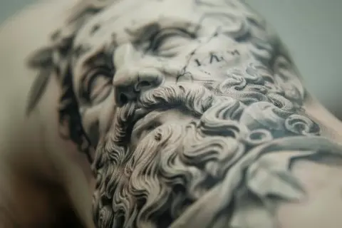 Detalle de un tatuaje de microrealismo conceptual de un rostro clásico con barba rizada y corona de laurel.