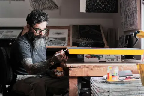 Thomas Hooper, con barba y gafas verdes, pinta un objeto amarillo en su estudio.