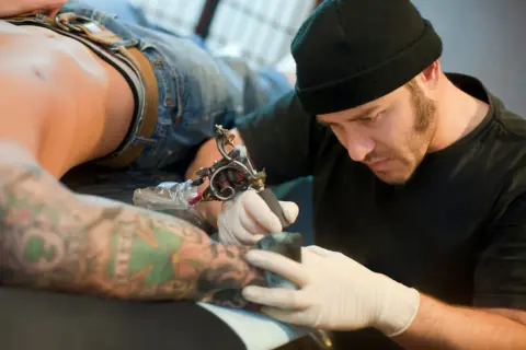 Tatuador con gorro negro y guantes blancos tatuando el brazo de un cliente con tatuajes coloridos.