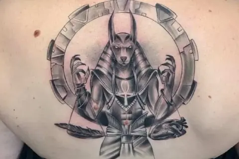 Tatuaje de Anubis con balanza, pluma y corazón, rodeado por un círculo mecánico.