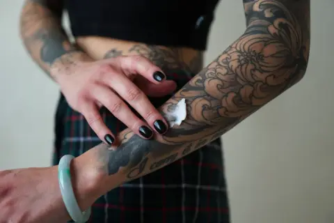Mano con uñas negras aplicando crema sobre un tatuaje de estilo dark work en el brazo, con diseño de olas y flores.