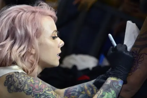 Artista de tatuajes con cabello rosa, con un diseño de megan massacre en su brazo, mientras otro artista trabaja en su piel.