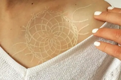 Un delicado tatuaje white out con diseño de atrapasueños en la clavícula.