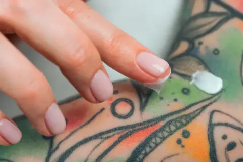 Dedos aplicando crema sobre un tatuaje colorido, parte de un proceso de cuidado y tatuaje terapéutico.
