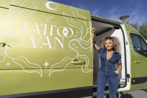 Una mujer sonriente posa junto a una furgoneta verde decorada con "The Tattoo Van". ¡Un estudio de tatuajes camper listo para viajar!