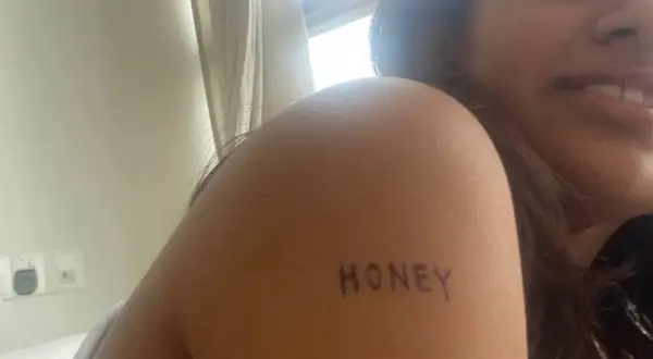 Un delicado tatuaje handpoke con la palabra "HONEY" en el brazo de una mujer sonriente.