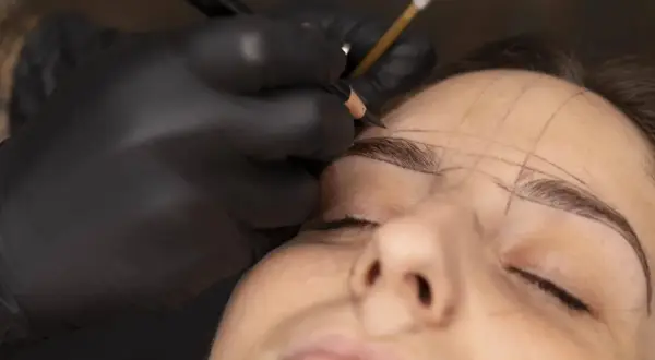 Manos enguantadas dibujan cejas con lápiz, marcando el rostro de Jessica Wilde para un diseño preciso.