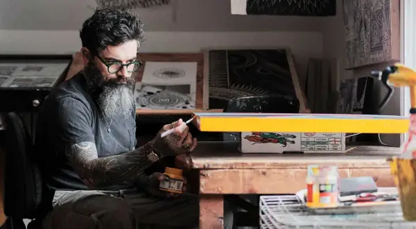 Thomas Hooper, con barba y gafas verdes, pinta un objeto amarillo en su estudio.