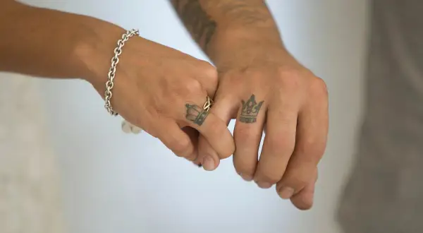 Tatuaje manos entrelazadas significado: dos coronas en los dedos, símbolo de unión y realeza compartida.