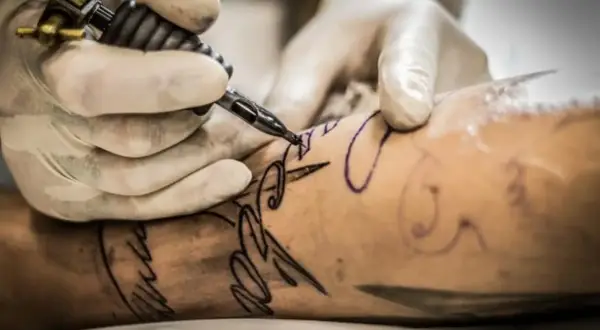 Manos enguantadas crean un diseño de letra con una máquina de tatuar. Un tatuaje con buena tinta se está formando en la piel.