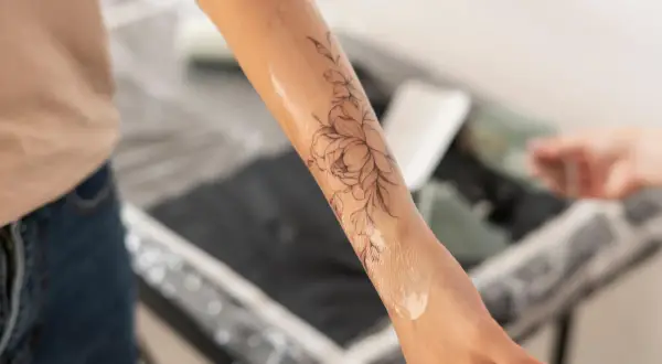 Brazo con un diseño de flores en proceso de tatuaje. El artista aplica una loción, similar a los cuidados post-tatuajes médicos, para hidratar la piel.