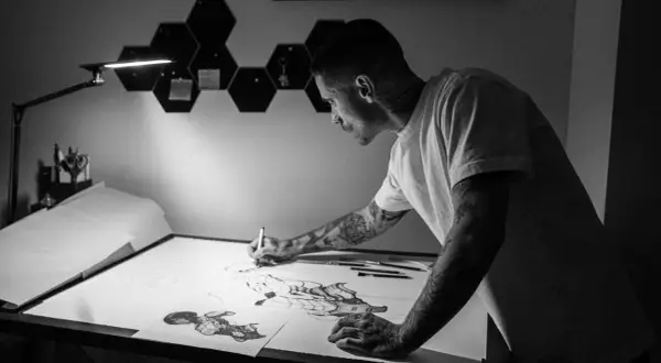 Víctor del Fueyo, artista con tatuajes, dibuja con concentración bajo una lámpara.