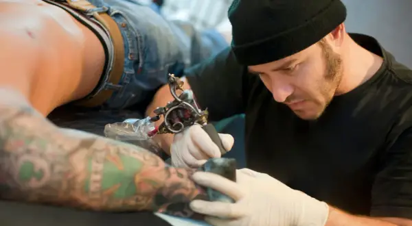 Tatuador con gorro negro y guantes blancos tatuando el brazo de un cliente con tatuajes coloridos.