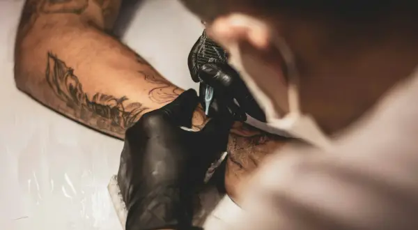 Artista con guantes negros usa máquina para crear un tatuaje en un brazo con diseños previos. La emla para tatuajes ayuda a que el proceso sea más cómodo.
