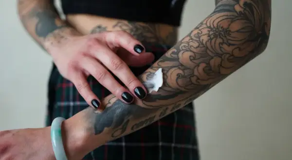 Mano con uñas negras aplicando crema sobre un tatuaje de estilo dark work en el brazo, con diseño de olas y flores.