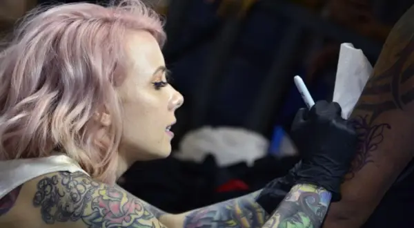 Artista de tatuajes con cabello rosa, con un diseño de megan massacre en su brazo, mientras otro artista trabaja en su piel.
