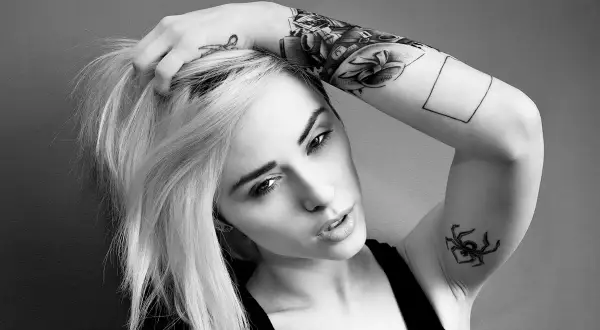 Alysha Nett, con su cabello rubio y tatuajes llamativos, posa con una mano en su cabeza.