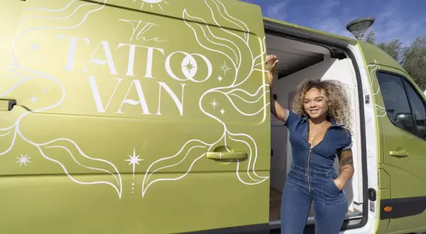Una mujer sonriente posa junto a una furgoneta verde decorada con "The Tattoo Van". ¡Un estudio de tatuajes camper listo para viajar!