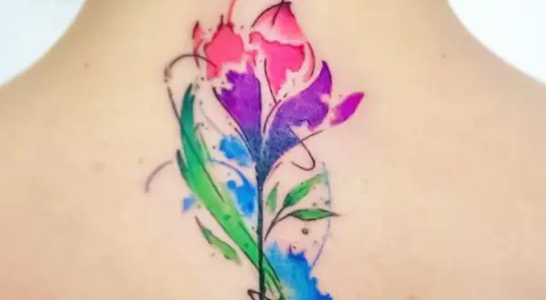 Un vibrante tatuaje de flor estilo acuarela en la espalda, con tonos rosas, morados y verdes. Un diseño de tatuajes de colores para mujer que evoca la naturaleza.