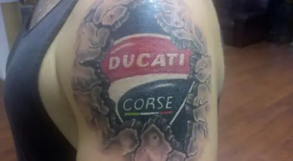 Tatuaje de un logo de Ducati Corse en el hombro, con efecto de piel rasgada.