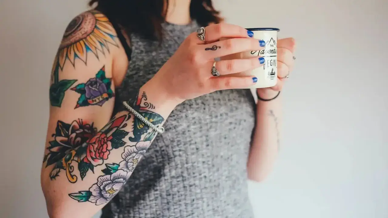 Brazos llenos de tatuajes coloridos, como arreglar un tatuaje mal hecho. Una mujer sostiene una taza.