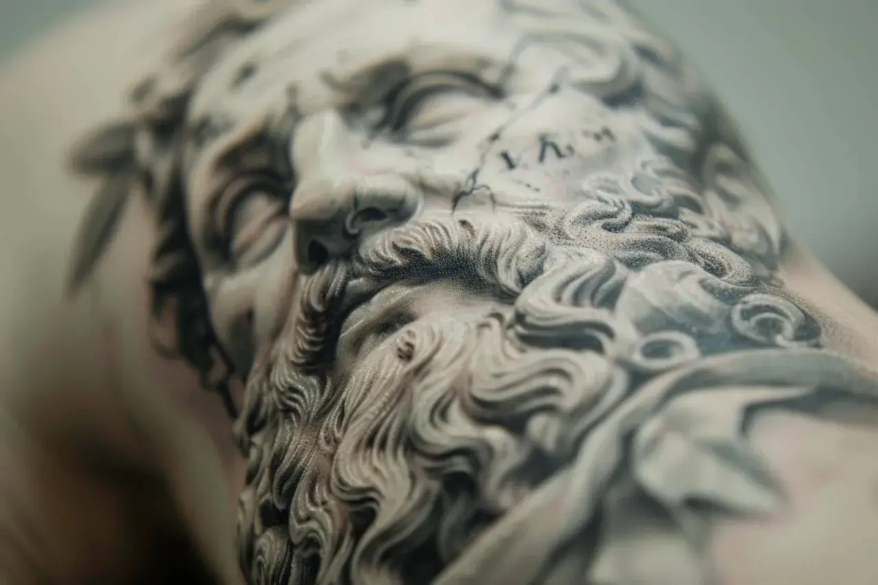 Detalle de un tatuaje de microrealismo conceptual de un rostro clásico con barba rizada y corona de laurel.