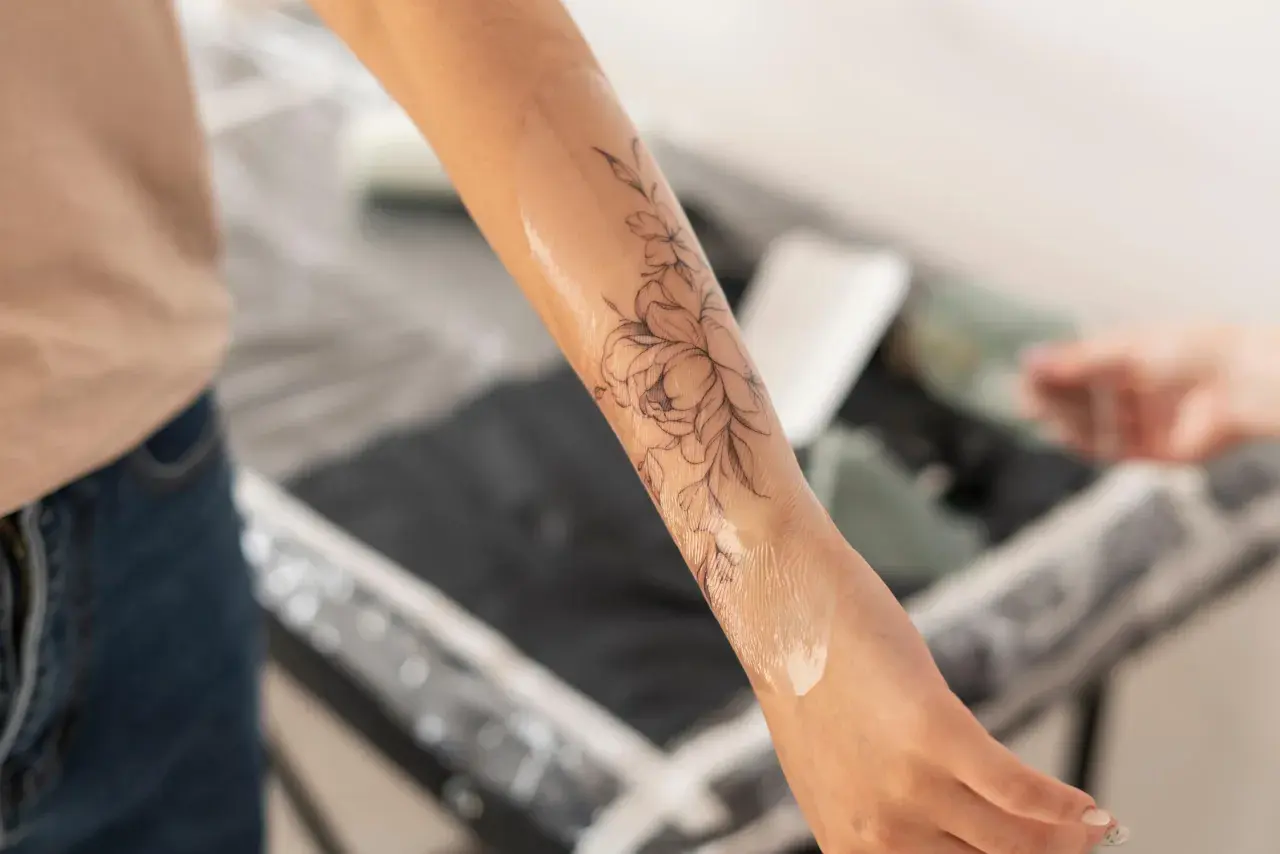 Brazo con un diseño de flores en proceso de tatuaje. El artista aplica una loción, similar a los cuidados post-tatuajes médicos, para hidratar la piel.