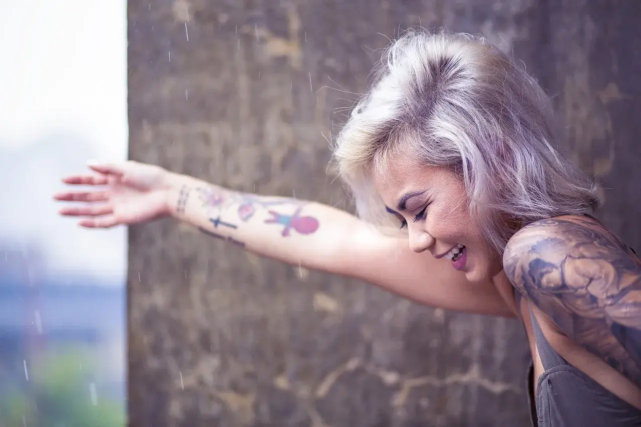 Mujer sonríe bajo la lluvia, con su brazo lleno de tatuajes, recordando el día que decidió hacerse un tatuaje.