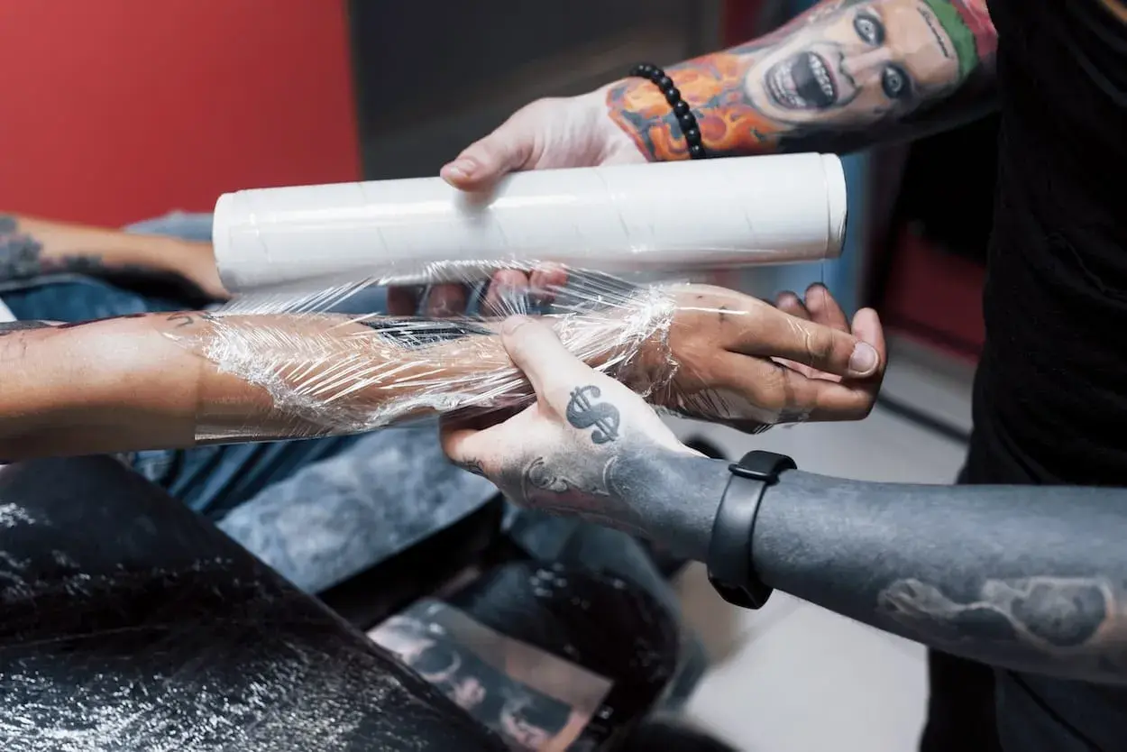 Man wrapping a fresh tattoo with plastic wrap. La curación es clave, y te preguntarás cuántos días hay que ponerse crema en un tatuaje para que quede perfecto.