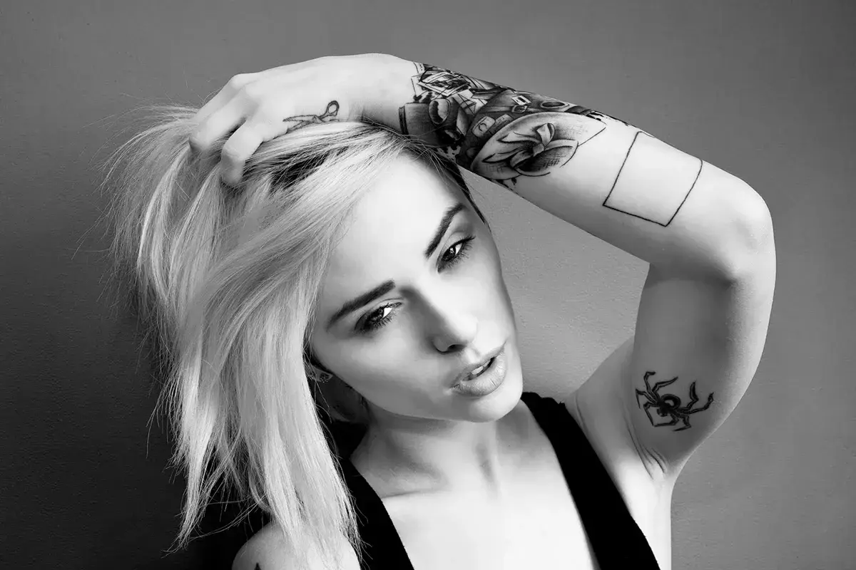 Alysha Nett, con su cabello rubio y tatuajes llamativos, posa con una mano en su cabeza.