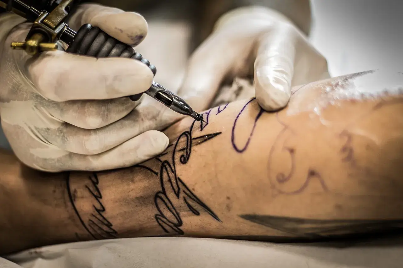 Manos enguantadas trabajan con una máquina de tatuar sobre un brazo, delineando un diseño. Aprende como cuidar un tatuaje recien hecho.