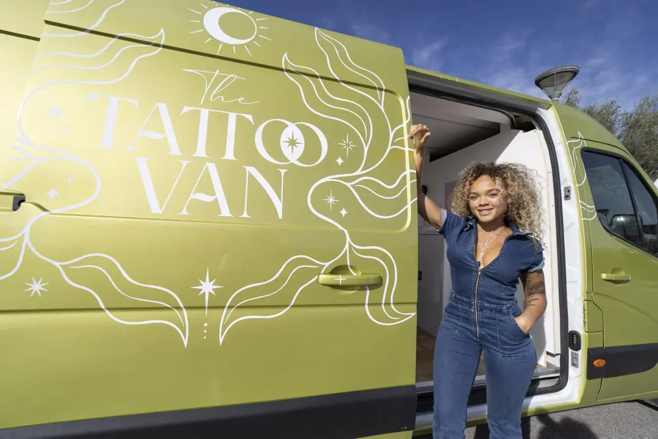 Una mujer sonriente posa junto a una furgoneta verde decorada con "The Tattoo Van". ¡Un estudio de tatuajes camper listo para viajar!