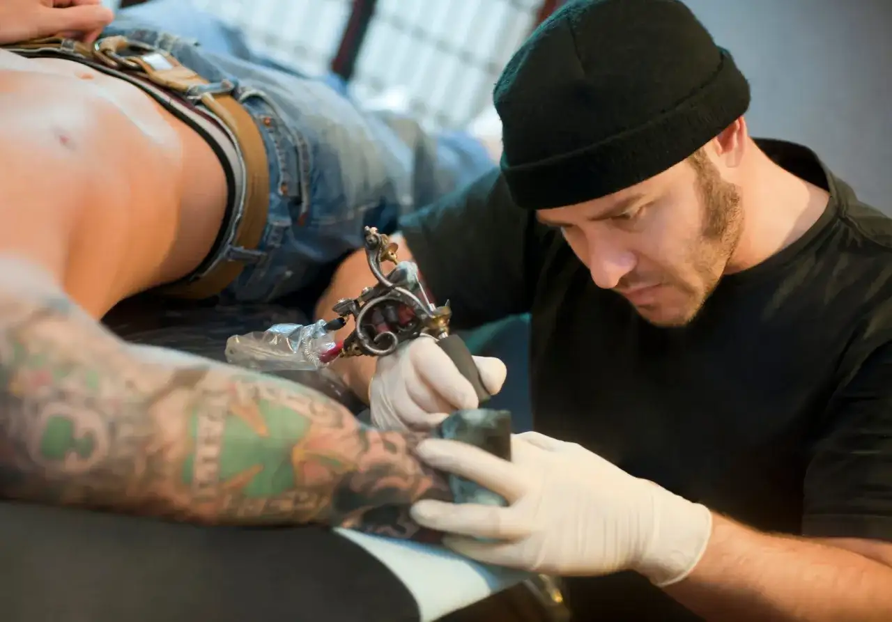 Tatuador con gorro negro y guantes blancos tatuando el brazo de un cliente con tatuajes coloridos.