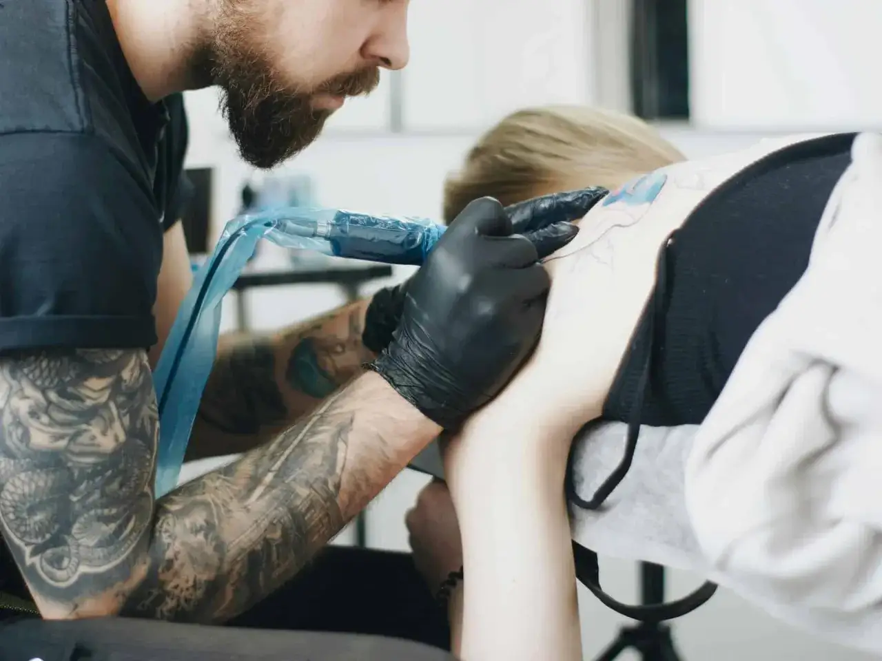 Artista con barba y tatuajes empieza a tatuar en el hombro de una persona.