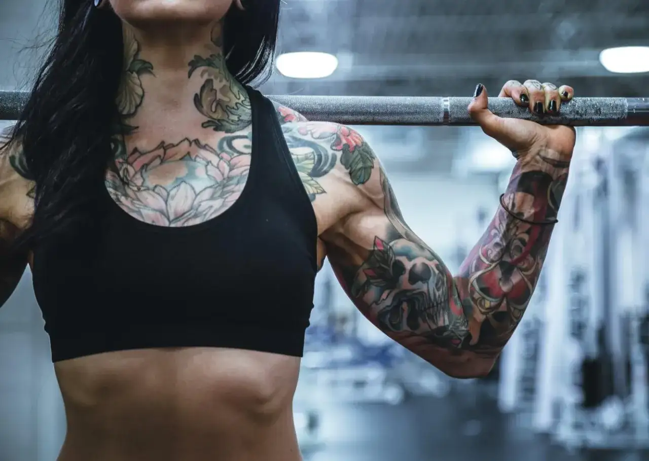 Mujer con un impresionante tatuaje en el cuello y brazos, levantando una barra. Su cuerpo fuerte muestra la dedicaci&oacute;n al deporte y al arte corporal.