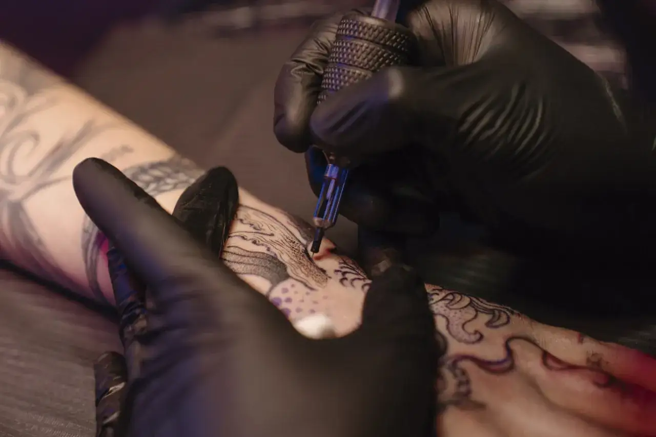 Manos enguantadas trabajando en un tatuaje intrincado. El artista usa una máquina de tatuar para aplicar buena tinta tattoo en la piel, creando un diseño detallado.