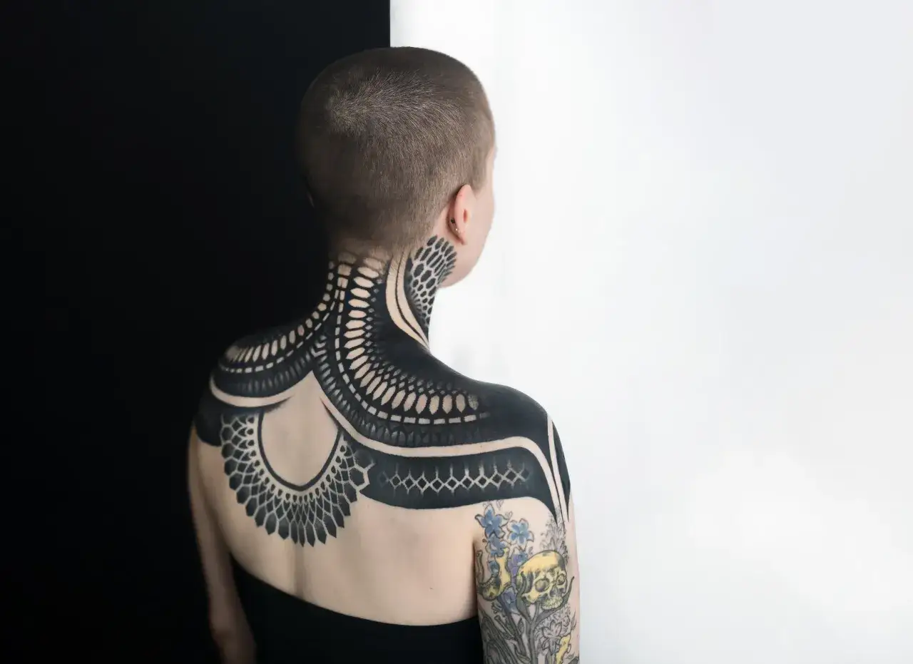 Una persona con un intrincado tattoo negro que cubre su cuello, hombros y espalda. El dise&ntilde;o es geom&eacute;trico y org&aacute;nico, con detalles que recuerdan a escamas o plumas.